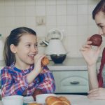 Happy young mother and cute daughter having breakfast eating muffins and talking at home in modern kitchen. Family, food, home and people concept